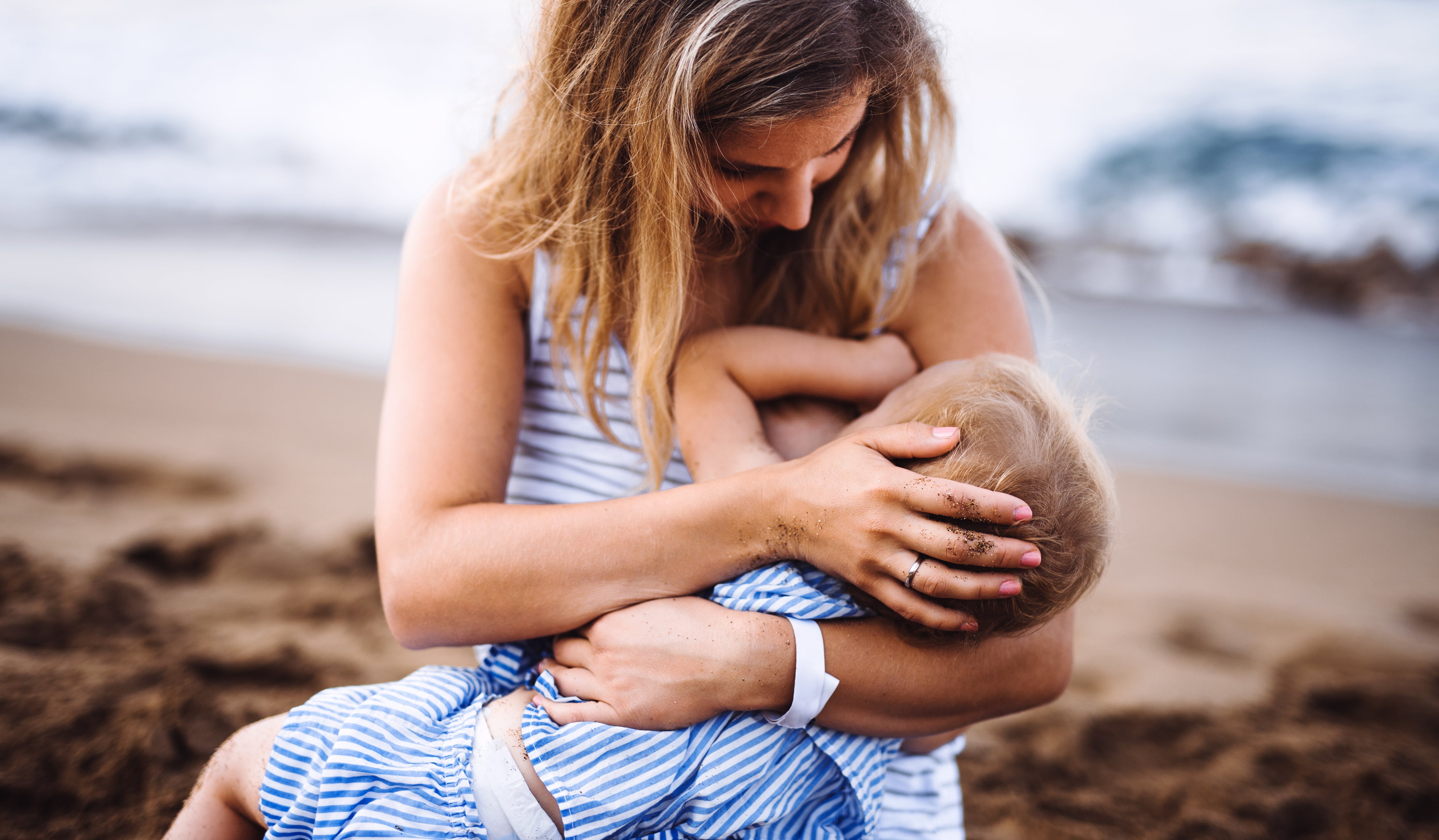 Young mother breasfeeding toddler daughter on beach on summer holiday.