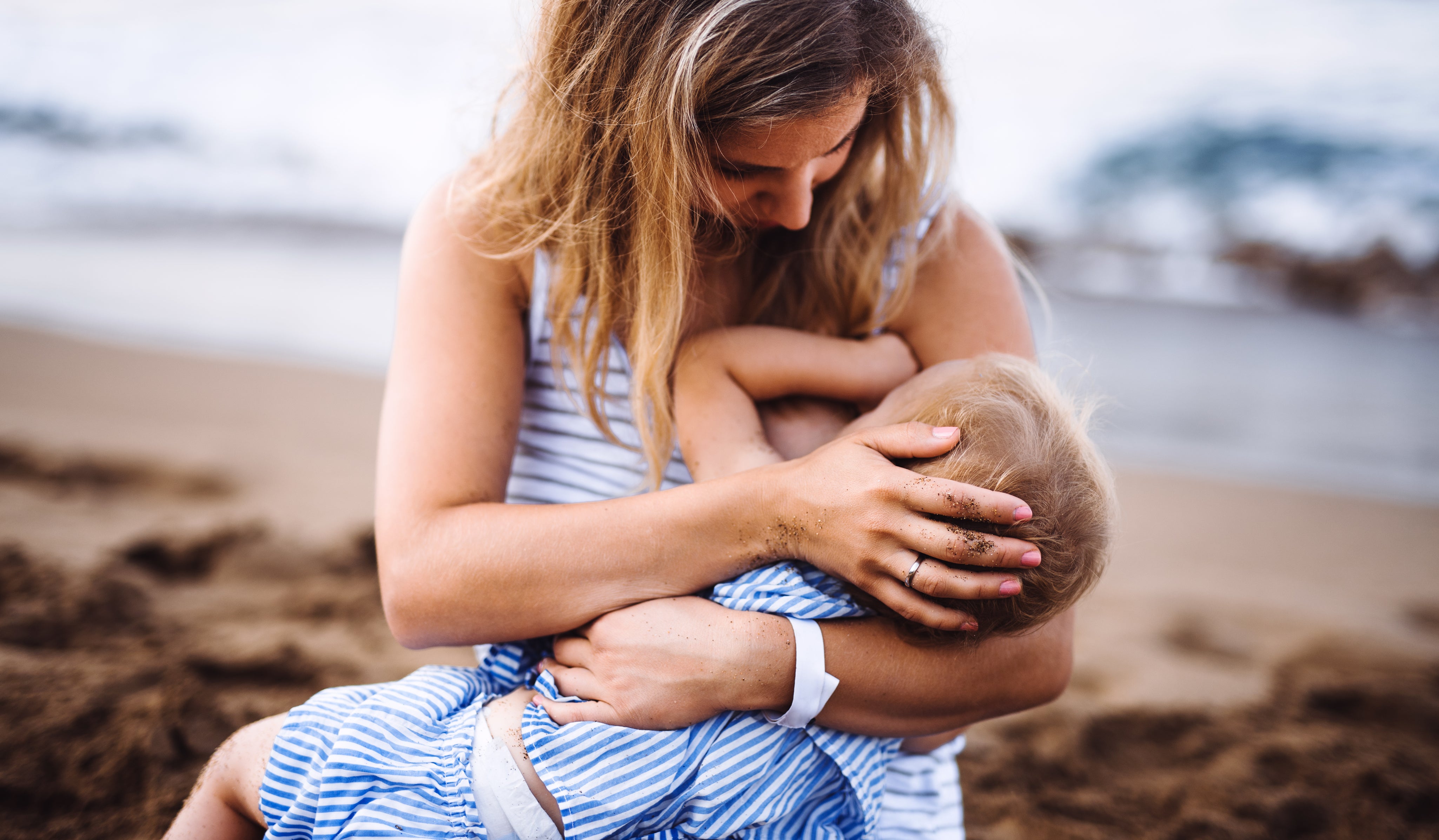 Young mother breasfeeding toddler daughter on beach on summer holiday.
